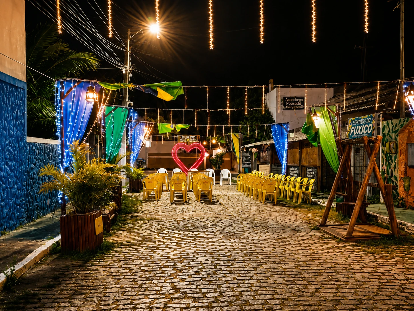 Rua do Fuxico à noite em Genipabu, iluminada por cordões de luzes, com cadeiras amarelas e coração luminoso